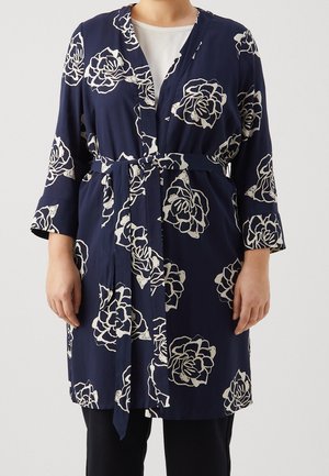 Granatowy płaszcz z paskiem i kremowym kwiatowym wzorem, noszony na białym topie i czarnych spodniach, pokazany od szyi do połowy uda.