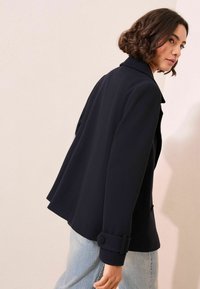 Femme aux cheveux courts et bouclés portant un manteau noir et un jean bleu clair, se retournant vers l'arrière devant un fond de mur pâle.
