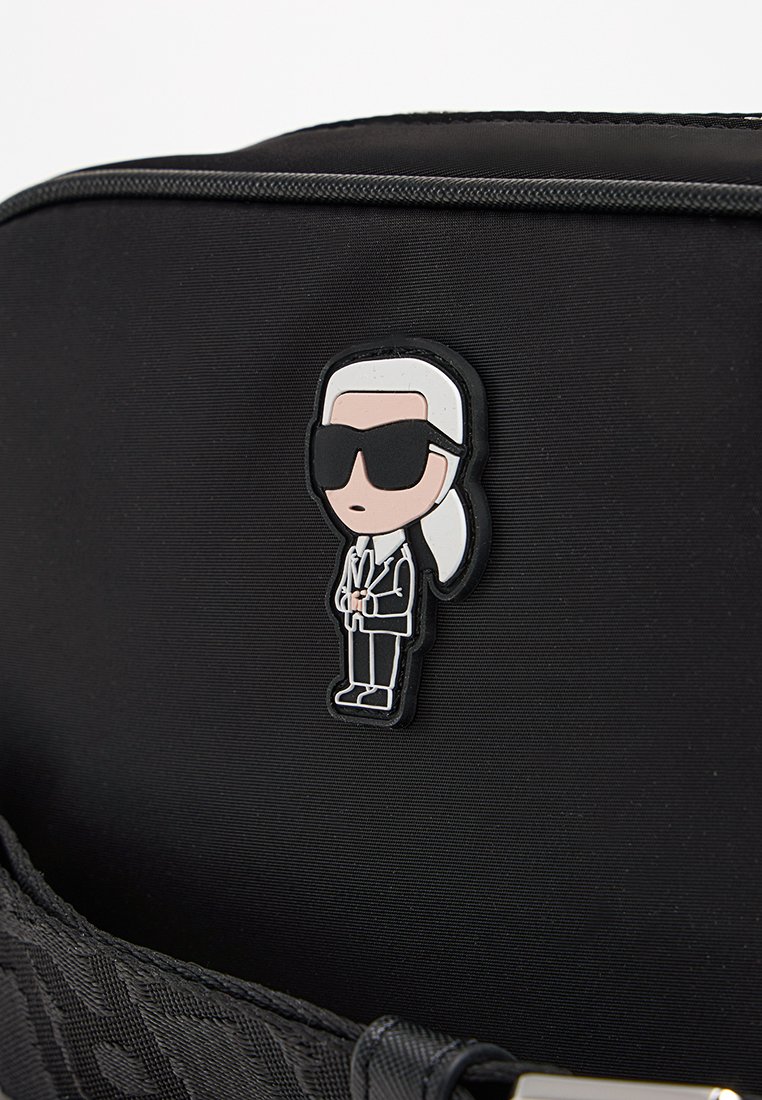 Bolsa de tela negra que presenta un pin de un personaje de dibujos animados de un hombre con gafas de sol y traje, con cabello blanco, adjunto a la superficie frontal.
