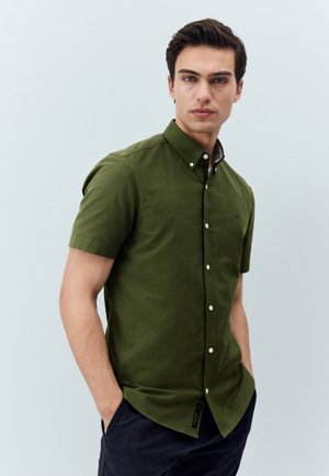 Mladi muškarac s tamnom kosom, obučen u maslinasto zelenu košulju kratkih rukava i tamne hlače, stoji s rukama u džepovima.