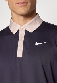 Tengerészkék póló világos rózsaszín gallérral, három gombos felső résszel és fehér Nike logóval a mellkasán. Sima textúrájú és lélegző anyagú.
