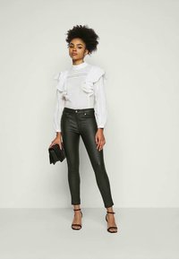 Blusa branca com detalhes em babados, mangas longas rendadas, combinada com calças justas de couro sintético pretas e sapatos de salto alto pretos com tiras. Bolsa de mão segurada.
