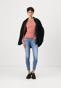 Maglietta rosa a maniche lunghe indossata sotto una camicia verde scuro, abbinata a jeans aderenti azzurri e scarpe nere, su uno sfondo neutro.