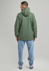 Green waterproof jacket with a hood, smooth texture, and long sleeves. Paired with light blue denim pants and white sneakers.