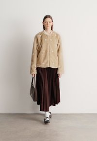 Casaco bege de pelo sintético com bordado floral, combinado com uma saia plissada castanho escuro. A modelo leva uma mala cinzenta e usa meias brancas com sapatos.