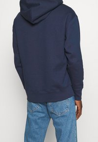 Sudadera azul marino, mezcla de algodón, con bolsillo canguro, puños y dobladillo acanalados, y con una capucha amplia. Lleva puestas unas jeans de mezclilla azul claro.