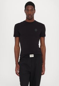 Tricou negru cu mâneci scurte, realizat dintr-un material neted. Prezintă o gâtă rotundă și un logo texturat pe piept. Design ajustat.