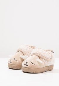 Chaussons en peluche beige avec un design de mouton, présentant des yeux brodés, des oreilles douces et une semelle texturée. Sangle ajustable pour un maintien sécurisé.