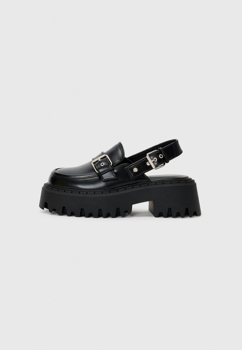 Mocassin à plateforme épaisse en cuir noir avec semelle crantée, détails de boucle argentée et bride arrière ouverte.