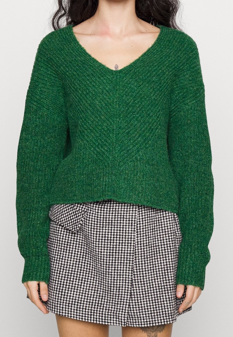 Zielony sweter dzianinowy z dekoltem w serek, z fakturowanym wzorem i opuszczonymi ramionami, zestawiony z czarno-białą spódnicą w kratę z kieszeniami.