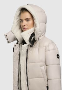 Nais elab beežis soojendusjakis, kus on suur fleece'i voodriga kapuuts ja must klamberdetail, vaatab külje poole ühetaolisel taustal.