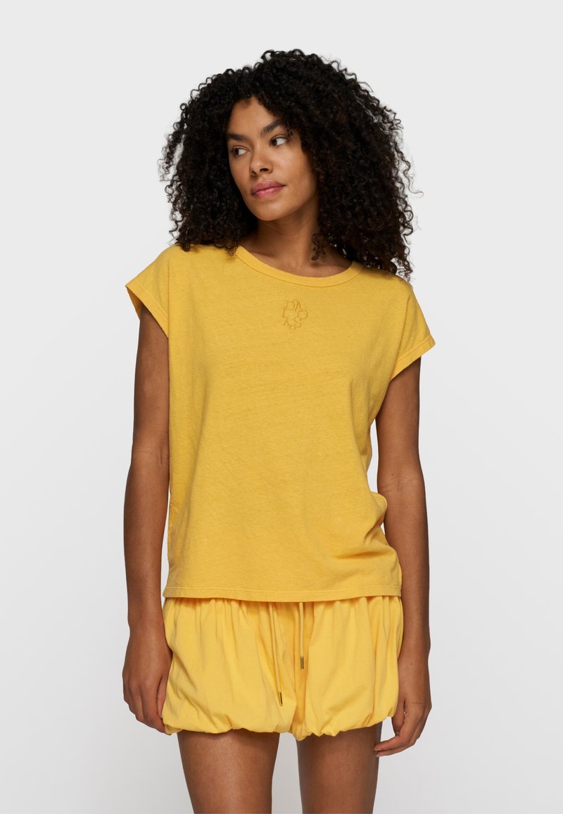 Mujer con cabello rizado, vestida con una blusa amarilla de manga corta y pantalones cortos amarillos holgados, de pie frente a un fondo liso de color gris claro.