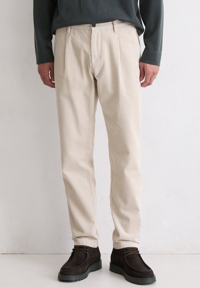Pantalon en velours côtelé beige avec une coupe décontractée, présentant des plis devant, une fermeture par bouton et un léger côtelage vertical, associé à des chaussures sombres.