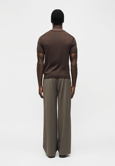 Άνδρας που έχει γυρισμένη την πλάτη του, φορά καφέ κοντομάνικο πουκάμισο και χαλαρά γκρι παντελόνια με φαρδύ πόδι, μπροστά σε απλό φόντο.