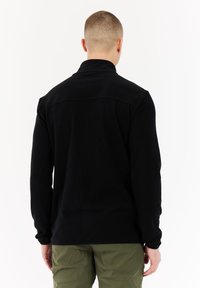 Uomo dai capelli corti che indossa una maglietta nera a maniche lunghe e pantaloni verde oliva, in piedi con la schiena rivolta verso l'osservatore su uno sfondo bianco.