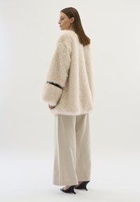 Faux-Fur-Mantel in Hellbeige mit lockerer Passform und weiten Ärmeln, versehen mit einem schwarzen Akzent an den Manschetten. Kombiniert mit cremefarbenen Hosen und Sch heels.