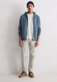 Blauer Hoodie mit Reißverschluss über einem weißen T-Shirt, hellbeige Hose und hellbraunes Schuhwerk. Glatter Stoff und lockere Passform mit Kapuze und Kordelzügen.