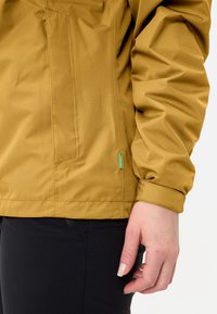 Brown textured jacket with a zippered pocket, elastic cuffs, and a green label accent. Worn over black pants.