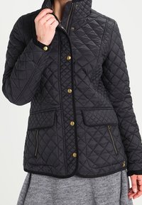 Quilted black jacket features a high collar, gold hardware buttons, two front pockets, and contrasting trim along the edges.