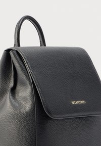 Mochila de cuero negro con acabado texturizado, cierre con solapa y asa superior. Detalles de logo en dorado en la parte frontal.