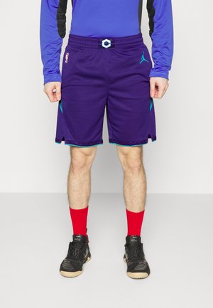 Purple basketball shorts with a drawstring waist, featuring teal accents, side pockets, and a logo on the left leg. Worn with red socks and black shoes.