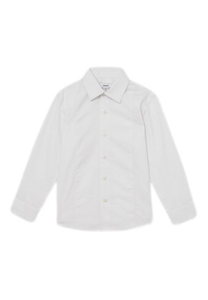 Niño joven que lleva una camisa blanca, pajarita negra y pantalones negros, de pie con una mano en el bolsillo contra un fondo liso.
