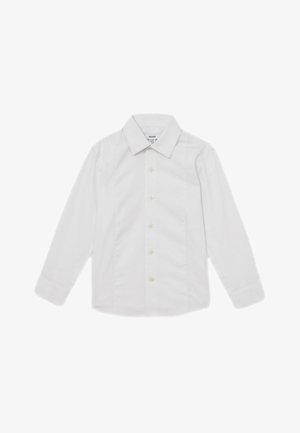 Niño joven que lleva una camisa blanca, pajarita negra y pantalones negros, de pie con una mano en el bolsillo contra un fondo liso.