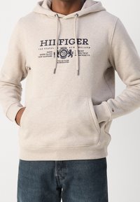 Sudadera color beige hecha de tela suave, con un bolsillo tipo canguro, capucha con cordón y un estampado gráfico en azul marino en la parte delantera. Puños y dobladillo de canalé.