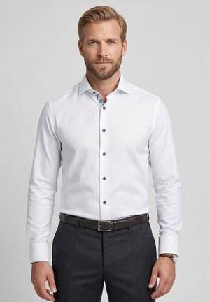 Homme blond portant une chemise blanche à boutons foncés, un pantalon sombre, une ceinture noire et une montre-bracelet marron.