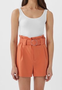 Un débardeur blanc à côtes avec un short orange taille haute doté d'une large ceinture et d'une boucle en métal circulaire. Le short est cintré et possède des poches latérales.