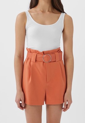 Un débardeur blanc à côtes avec un short orange taille haute doté d'une large ceinture et d'une boucle en métal circulaire. Le short est cintré et possède des poches latérales.