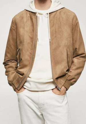 Homme portant une veste en daim brun clair par-dessus un sweat à capuche blanc et un pantalon blanc, les mains dans les poches, devant un fond uni.