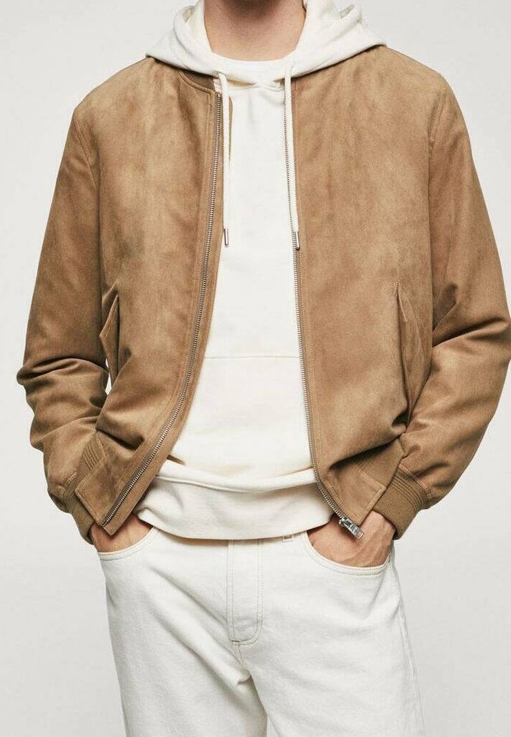 Homme portant une veste en daim brun clair par-dessus un sweat à capuche blanc et un pantalon blanc, les mains dans les poches, devant un fond uni.
