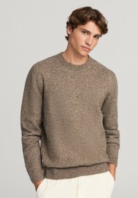 Maglione lavorato a maglia marrone chiaro con scollo rotondo, polsini e orlo a coste, caratterizzato da una finitura testurizzata. Indossato sopra pantaloni color crema.