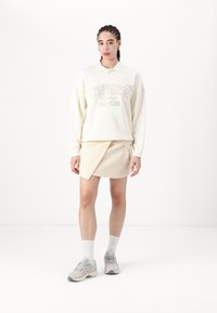 Sudadera de color beige claro con el logo "CONVERSE ALL-STAR", combinada con una falda beige a juego. La modelo lleva zapatillas grises y calcetas blancas.