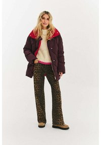 Pikowany bordowy płaszcz z czerwonym kołnierzem, beżowy sweter z dzianiny i brązowe spodnie w panterkę. Stylizację dopełniają beżowe buty.