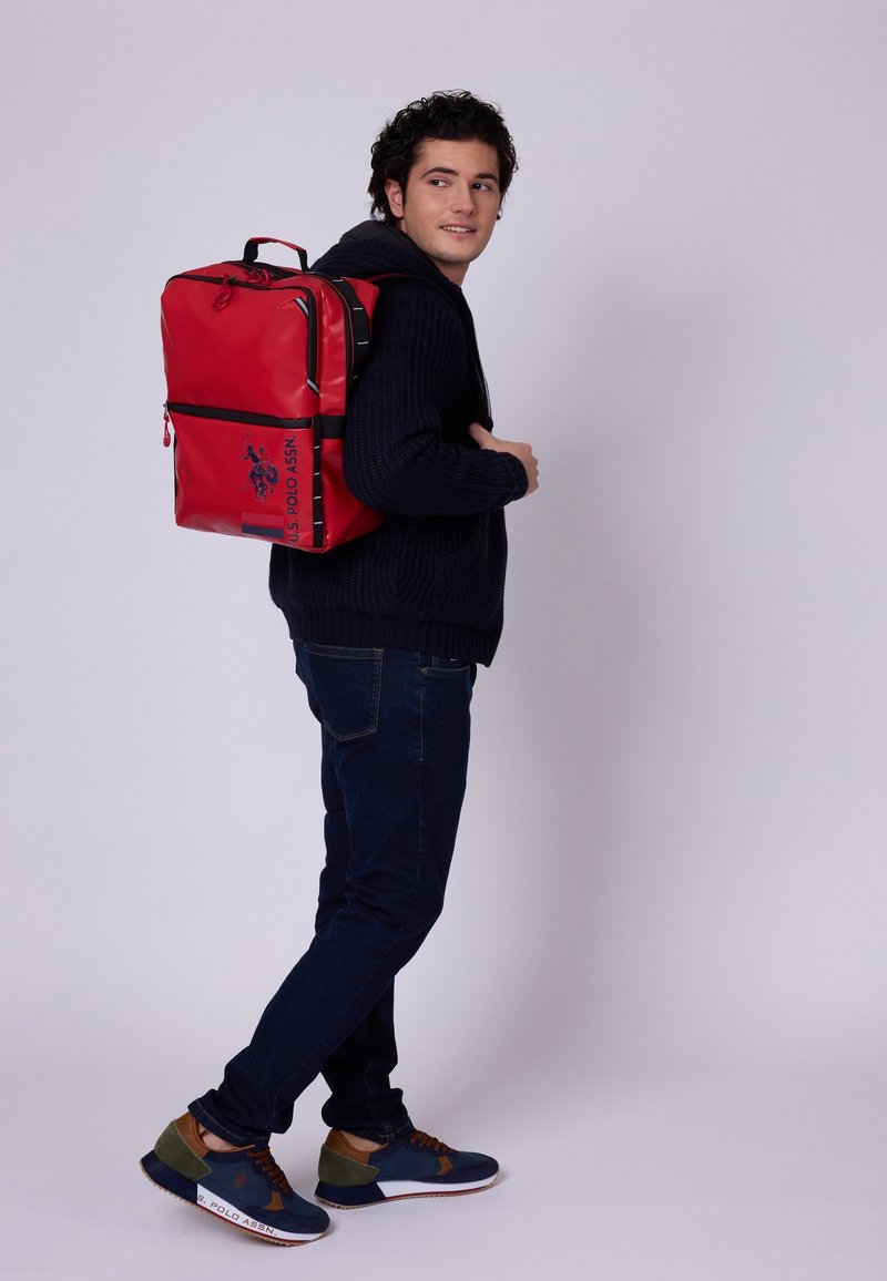 Sac à dos rouge en matériau durable, avec des accents et des zips noirs, de forme rectangulaire, et un logo sur le devant.