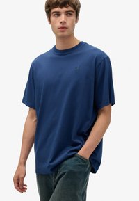 Camiseta de algodón azul marino con mangas cortas, cuello redondo, corte relajado y un pequeño logotipo en el pecho. Se combina con pantalones vaqueros de mezclilla.