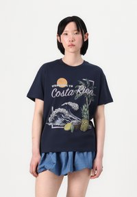 Navy cotton t-shirt featuring a graphic design with waves, palm trees, and fruits. Short sleeves and a round neckline. Paired with blue shorts.