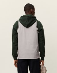 Hall mähkiv hall kapuutsiga jakke, millel on rohelised varrukad, tõmblukuga kapuuts, külje triibud ja mugav lõige. Hoides kätel mütsi.