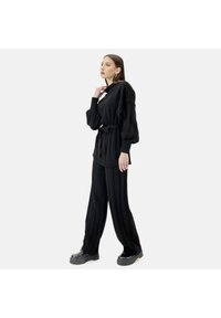 Conjunto de punto negro que presenta un suéter acanalado con cinturón, cuello alto y mangas anchas, combinado con pantalones de pierna ancha acanalados a juego y zapatos negros gruesos.