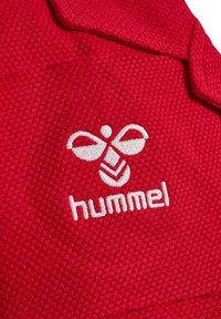 Czerwona, teksturowana tkanina z białym wyszywanym logo Hummel przedstawiającym stylizowaną trzmiela nad nazwą marki.