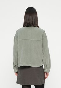 Donna con capelli scuri di lunghezza media indossa un giaccone verde in corduroy oversize e una mini gonna marrone a fantasia, vista da dietro.