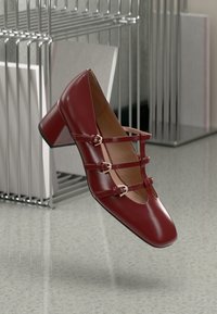 Red patent leather shoe with a square toe, mid-height block heel, and three decorative straps with buckles on the front.