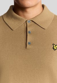 Tan poloträja i ribbat tyg, med krage, tre grå knappar och en liten gul broderad logotyp på sidan.