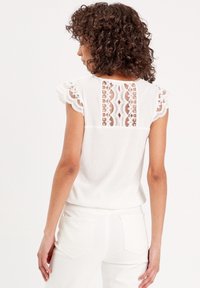 White blouse with short ruffled sleeves, textured fabric, and a lace back panel featuring a scalloped design. Paired with white pants.