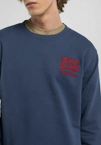 Felpa blu navy con collo e polsini a costine, dotata di un piccolo logo ricamato rosso sul petto. Tessuto morbido, vestibilità casual.