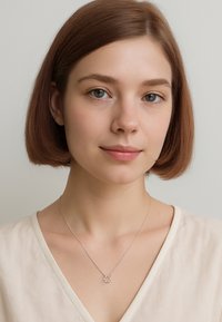 Sølvsmykke med cirkulært vedhæng, der har små sten, båret af en person med pagehår, mod en neutral baggrund.