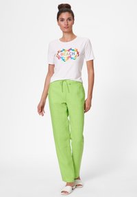 White t-shirt with "BEACH" graphic in colorful font, paired with loose green linen pants and white slide sandals, standing against a plain background.