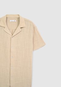 Camicia a maniche corte in beige, realizzata in un tessuto testurizzato, con colletto classico e cinque bottoni frontali, fondo dritto.